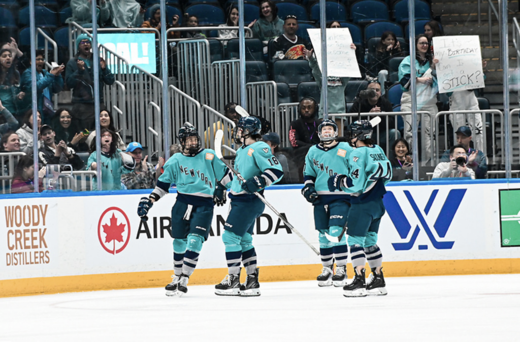 Four New York players, wearing teal, gather to celebrate a goal with a group hug.